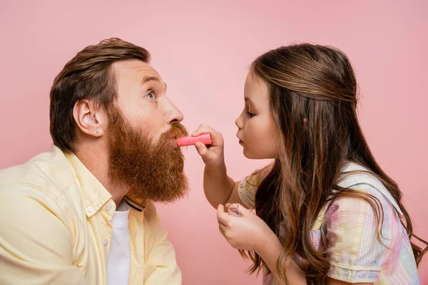Вид збоку дівчини, що наносить помаду на бородатого батька ізольовано на рожевому — стокове фото