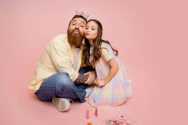 Bärtiger Mann mit Krone Haarband schmollen Lippen in der Nähe der Tochter und dekorative Kosmetik auf rosa Hintergrund — Stockfoto