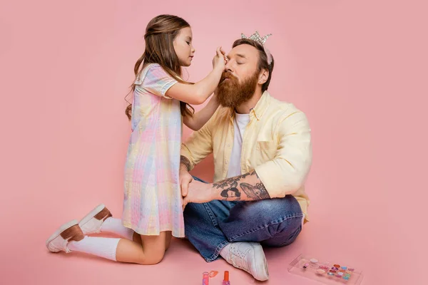Preteen girl applying decorative cosmetic on tattooed dad with crown headband on pink background — Stock Photo