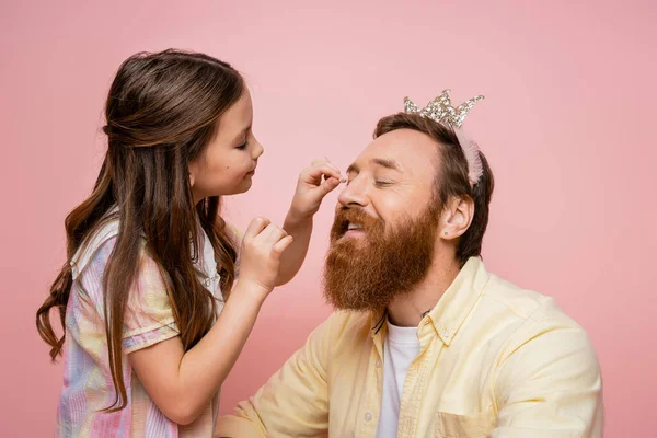 Sorrindo menina pré-adolescente aplicando sombra no pai barbudo com a cabeça da coroa isolada no rosa — Fotografia de Stock