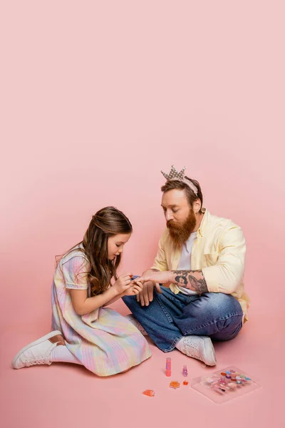 Menina pré-adolescente aplicando esmalte de unhas na mão de pai barbudo com cabeça de coroa no fundo rosa — Fotografia de Stock