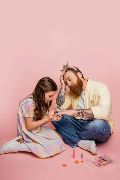 Niño preadolescente aplicando esmalte de uñas en la mano de papá molesto con diadema de corona sobre fondo rosa - foto de stock