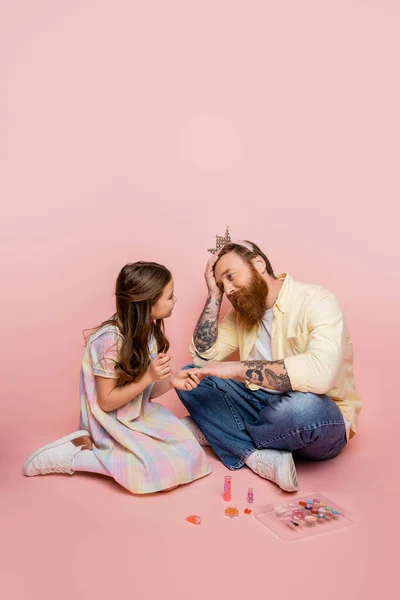 П'ятнадцять дівчат тримають лак для нігтів біля руки втомленого батька з коронкою на голові на рожевому фоні — стокове фото