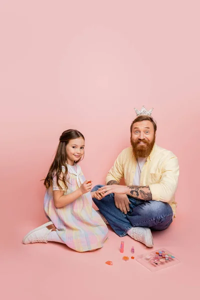 Веселая девушка держит лак для ногтей рядом с отцом с повязкой на голове и декоративной косметикой на розовом фоне — стоковое фото