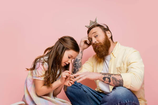 Preteen girl applying nail polish on hand of tattooed dad with crown headband on pink background — Stock Photo