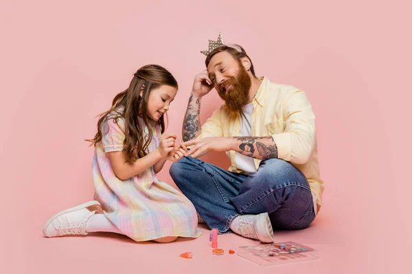Cheerful girl applying nail polish on hand of tattooed dad with crown headband on pink background — Stock Photo
