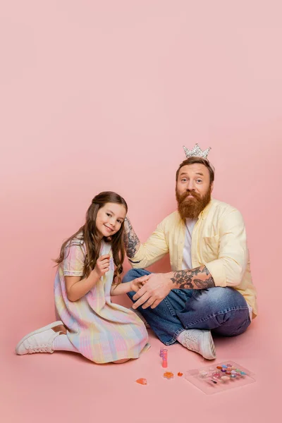Menina positiva segurando esmalte de unhas perto do pai tatuado com coroa na cabeça e cosméticos decorativos no fundo rosa — Fotografia de Stock