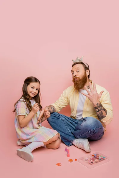 Улыбающийся ребенок держит лак для ногтей рядом с отцом с короной на голове дующиеся губы на розовом фоне — стоковое фото