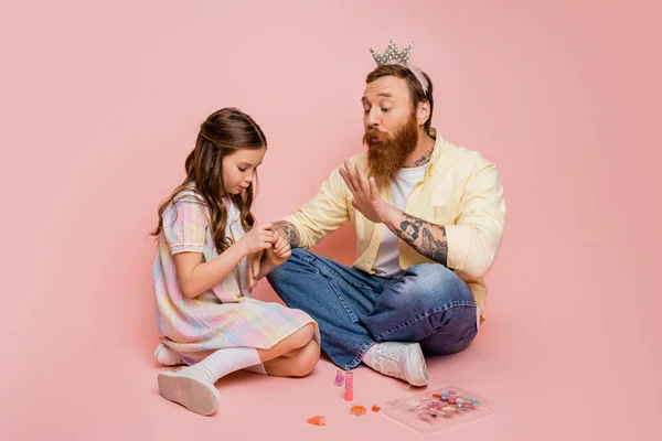 Homem com coroa na cabeça soprando na mão perto da filha com esmalte no fundo rosa — Fotografia de Stock