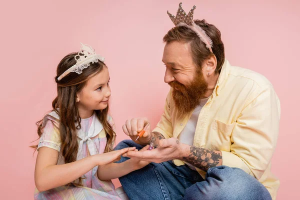 Hombre alegre con corona en la cabeza sosteniendo esmalte de uñas cerca de hija preadolescente sobre fondo rosa - foto de stock
