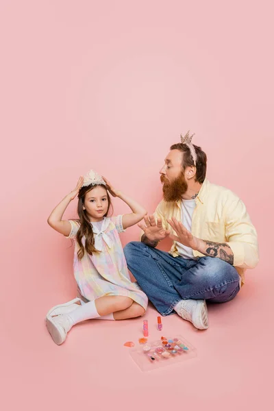 Papá barbudo con corona en la cabeza mirando a la hija preadolescente cerca de cosméticos decorativos sobre fondo rosa - foto de stock