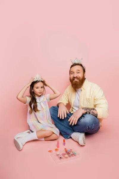 Hombre barbudo positivo mirando a la cámara cerca de la hija con corona y cosméticos decorativos sobre fondo rosa - foto de stock