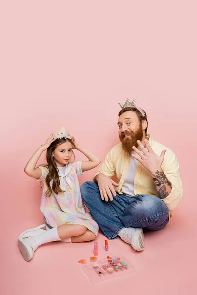 Preteen girl wearing crown near bearded father and decorative cosmetics on pink background — Stock Photo