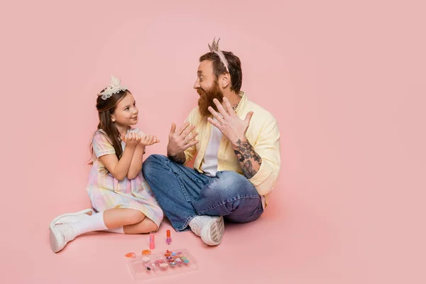 Alegre padre e hija con diademas de corona sentados cerca de cosméticos decorativos sobre fondo rosa - foto de stock