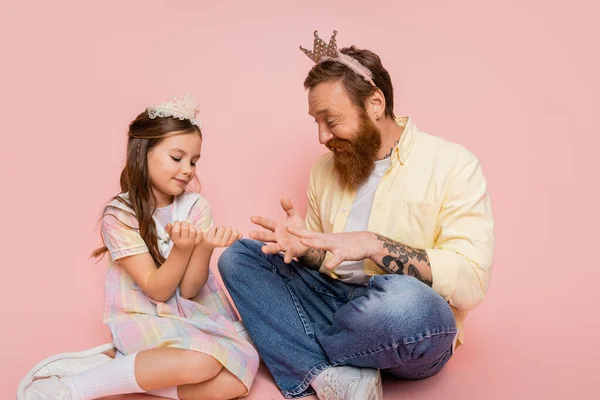 Overjoyed man with crown headband looking at hands of daughter on pink background — Stock Photo