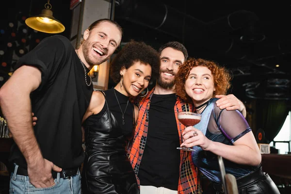 Positive redhead woman holding foam cocktail near interracial friends looking at camera and hugging in bar — Stock Photo