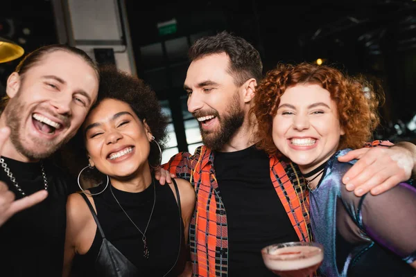 Веселий бородатий чоловік обіймає міжрасових друзів з коктейлем, розважаючись в барі — стокове фото