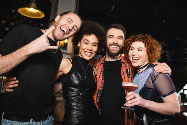 Веселая рыжая женщина с коктейлем рядом с многонациональными друзьями смотрит в камеру в баре — стоковое фото