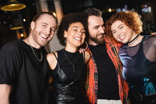 Bearded man hugging multiethnic friends and smiling at camera in bar — Stock Photo