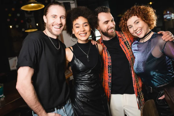 Cheerful multiethnic friends looking at camera and hugging in bar at night — Stock Photo