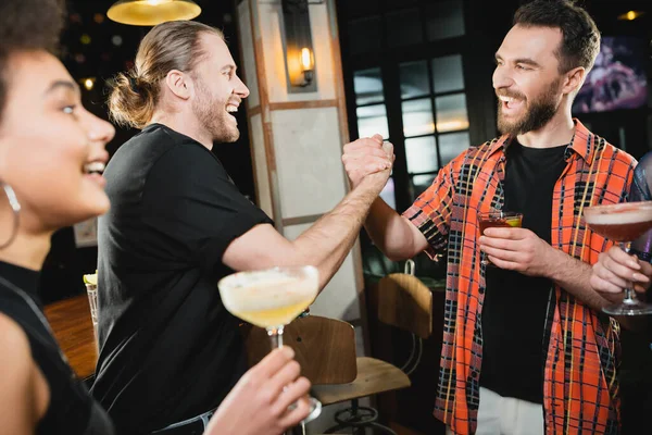 Cheerful bearded friends with cocktails holding hands while meeting in bar — Stock Photo
