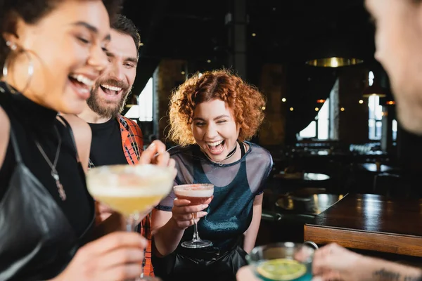 Femme rousse insouciante passer du temps avec des amis interracial tenant des cocktails dans le bar — Photo de stock