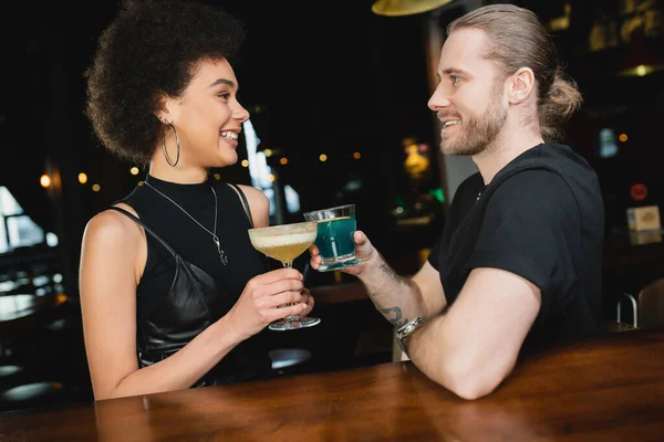Side view of positive man holding blue lagoon cocktail near african american friend in bar — Stock Photo