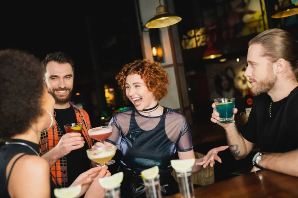 Aufgeregt lockige Frau im Gespräch mit afrikanisch-amerikanischen Freund mit Cocktail in der Nähe von Männern in Bar — Stockfoto