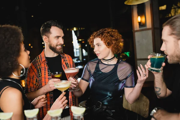 Multiethnische Freunde halten Cocktails in der Hand und unterhalten sich in der Nähe verschwommener Tequila-Gläser in einer Bar — Stockfoto