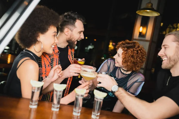 Vista lateral de amigos multiétnicos con diferentes cócteles hablando cerca borrosa vasos de tequila en el bar - foto de stock