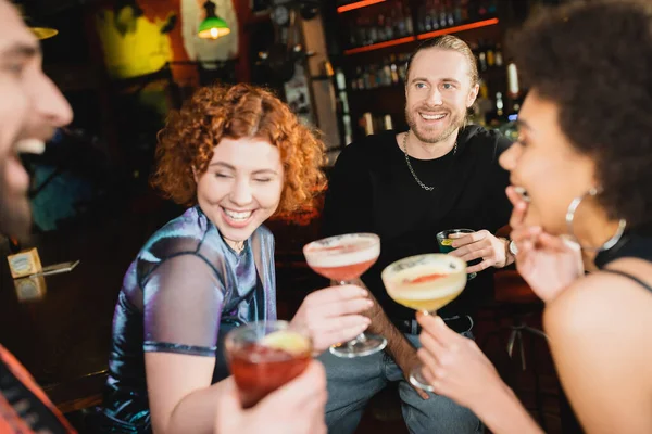 Positive man holding blue lagoon cocktail near interracial friends talking in bar — Stock Photo