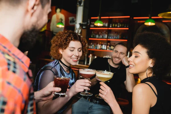 Улыбающиеся многонациональные женщины, звенящие коктейлями возле размытых друзей в баре — стоковое фото