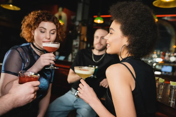 Positivo encaracolado afro-americano mulher segurando pisco coquetel azedo perto de amigos no bar — Fotografia de Stock