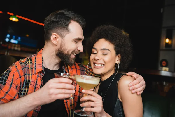 Hombre positivo sosteniendo negroni cóctel y abrazando joven afroamericano amigo en el bar - foto de stock