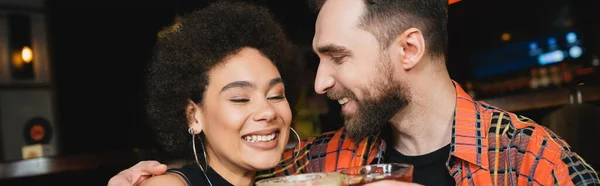 Веселый мужчина обнимает молодого африканского друга с коктейлем в баре, баннер — стоковое фото