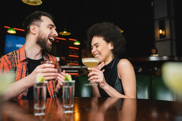 Возбужденные межрасовые друзья, держащие коктейли и смеющиеся возле размытых рюмок текилы в баре — стоковое фото