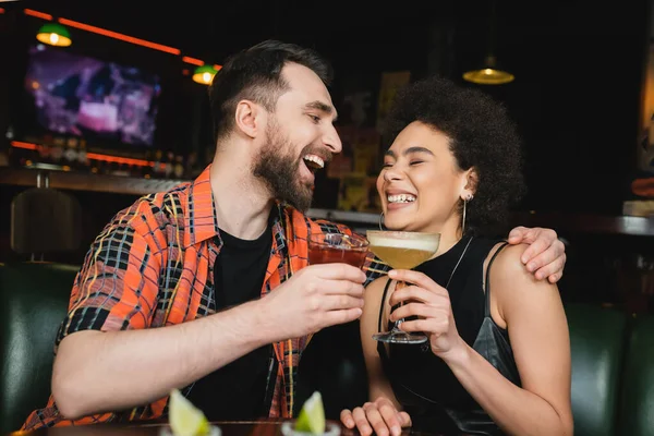 Laughing man holding negnoni cocktail and hugging african american friend near blurred tequila in bar — Stock Photo