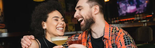 Cheerful man hugging african american friend with cocktail in bar, banner — Stock Photo