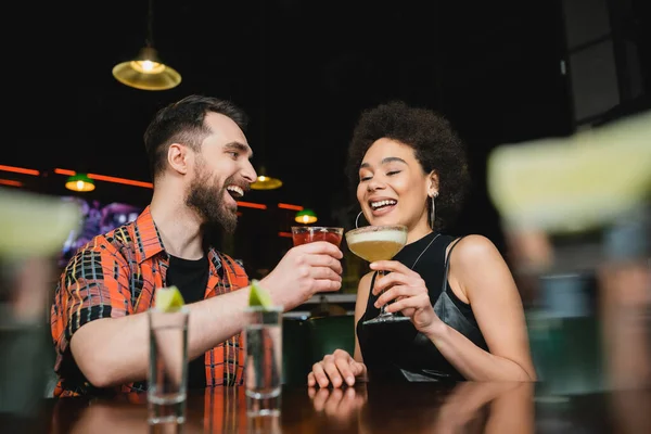 Overjoyed interracial friends holding cocktails near tequila shots on stand in bar — Stock Photo