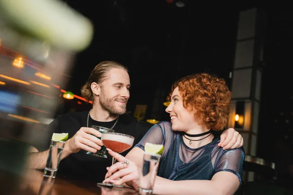 Позитивний чоловік обіймає червонуватого друга з коктейлем біля підставки в барі — стокове фото