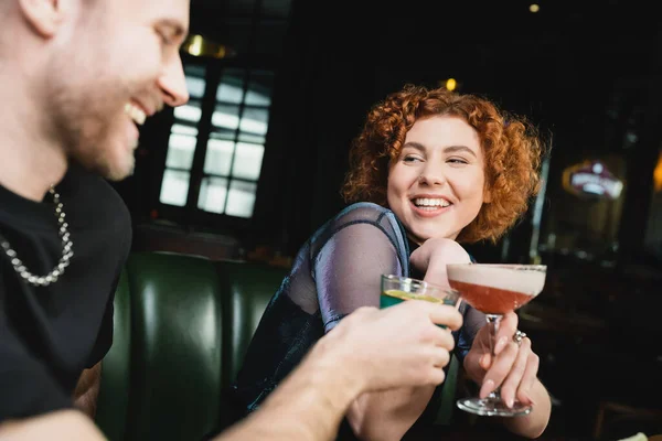Positive redhead woman clinking clover club cocktail with blurred friend in bar — Stock Photo