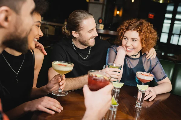 Smiling multiethnic friends hugging and holding cocktails near tequila shots in bar — Stock Photo