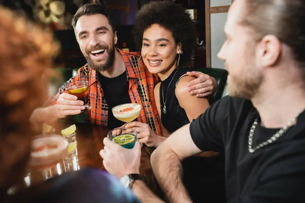 Personas multiétnicas positivas celebrando diferentes cócteles cerca de amigos borrosos en el bar - foto de stock