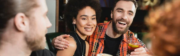 Усміхнений чоловік тримає негритянський коктейль і обіймає афроамериканського друга в барі ввечері, банер — стокове фото