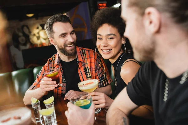 Alegre amigos inter-raciais falando perto de coquetéis e tiros de tequila com limão fresco no bar — Fotografia de Stock