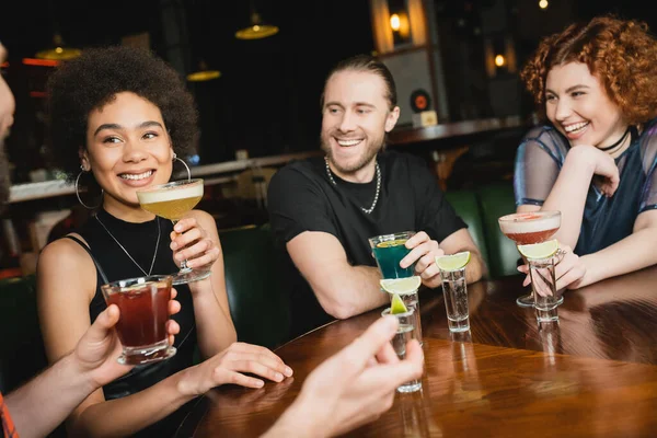 Femme afro-américaine joyeuse tenant un cocktail et regardant un ami flou dans le bar — Photo de stock