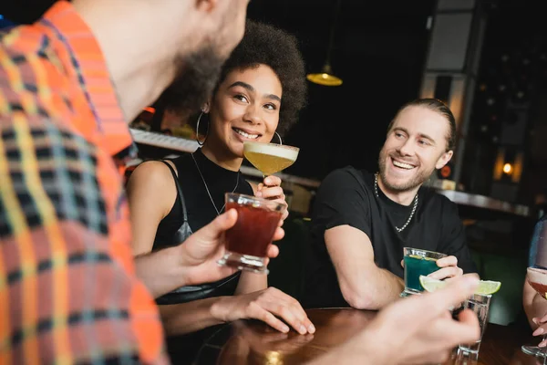 Улыбающаяся африканская американка, держащая коктейль писко сауэр рядом с другом в баре ночью — стоковое фото