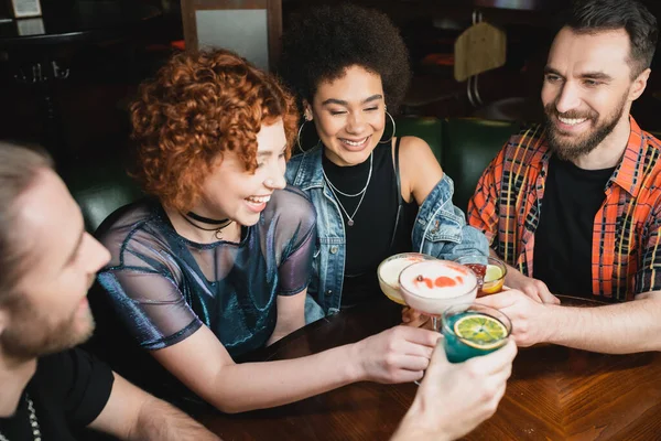 Усміхнені міжрасові жінки тримають коктейлі біля бородатих друзів в барі — стокове фото
