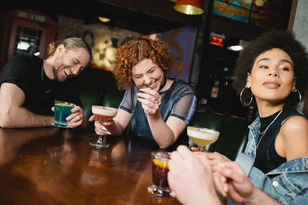 Happy interracial friends laughing and holding different cold cocktails in bar — Stock Photo