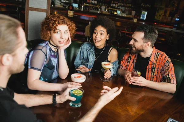 Des personnes multiethniques excitées regardant un ami tout en parlant à proximité de différents cocktails dans le bar — Photo de stock